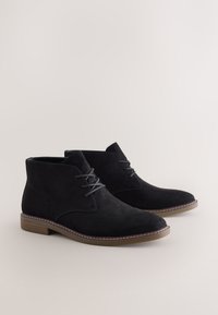 Bottines en daim noir à bout rond, lacets à l'avant et semelle en caoutchouc marron contrastante, avec des accents de couture subtils.
