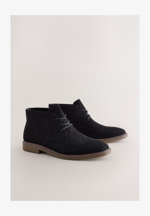 Black suede ankle boots with a rounded toe, lace-up front, and a contrasting brown rubber sole, featuring subtle stitching accents.