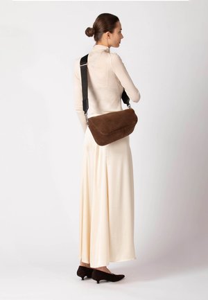 Braune Wildledertasche mit geschwungener Form, schwarzem verstellbarem Riemen und silberfarbenen Beschlägen. Quer getragen, kombiniert mit einem hellbeigen Kleid.