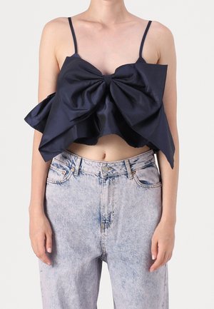 Kobieta nosząca granatowy crop top z dużą kokardą i cienkimi ramiączkami, w połączeniu z jasnoniebieskimi jeansami z wysokim stanem.