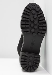 Semelle de chaussure noire avec une bande de roulement profonde à crampons, motifs hexagonaux et rectangulaires pour l'adhérence, inclinée contre un mur blanc et un sol.