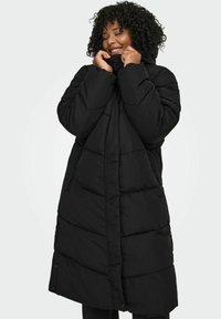 Melns pūkains mētelis ar garu, šūtu dizainu, rāvējslēdzēju un sprādzēm, kapuci un sānu kabatām, kas nodrošina praktiskumu un siltumu.