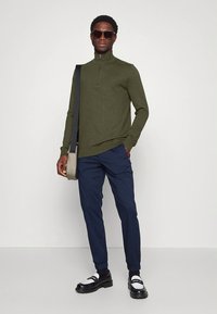 Pulover cu fermoar la gât, culoare verde măsliniu, pantaloni bleumarin, pantofi alb-negru, ochelari de soare, ținând o geantă ușoară de mesager.