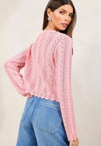 Rosa gestrickter Pullover mit Zopfmuster, knöchellang, mit geripptem Saum und langen Ärmeln, kombiniert mit hoch taillierten blauen Jeans.