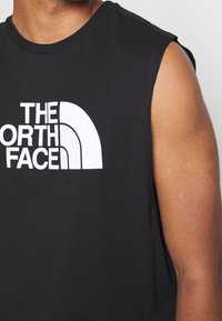 Haut du corps d’une personne portant un t-shirt noir sans manches avec le logo blanc The North Face sur la poitrine.