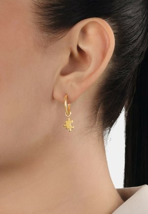Orecchini a forma di pezzo di puzzle in oro con una finitura lucida, caratterizzati da un piccolo design pendente e un attacco ad anello, esposti su un orecchio.