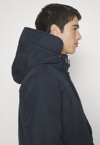 Navy zip-up jacket with a high collar and adjustable hood, featuring a textured fabric and side pockets.