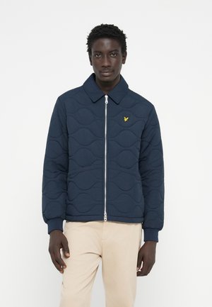 Veste matelassée marine avec fermeture éclair, dotée d'un col évasé et d'un petit logo jaune. Texture douce avec un motif ondulé sur le tissu.