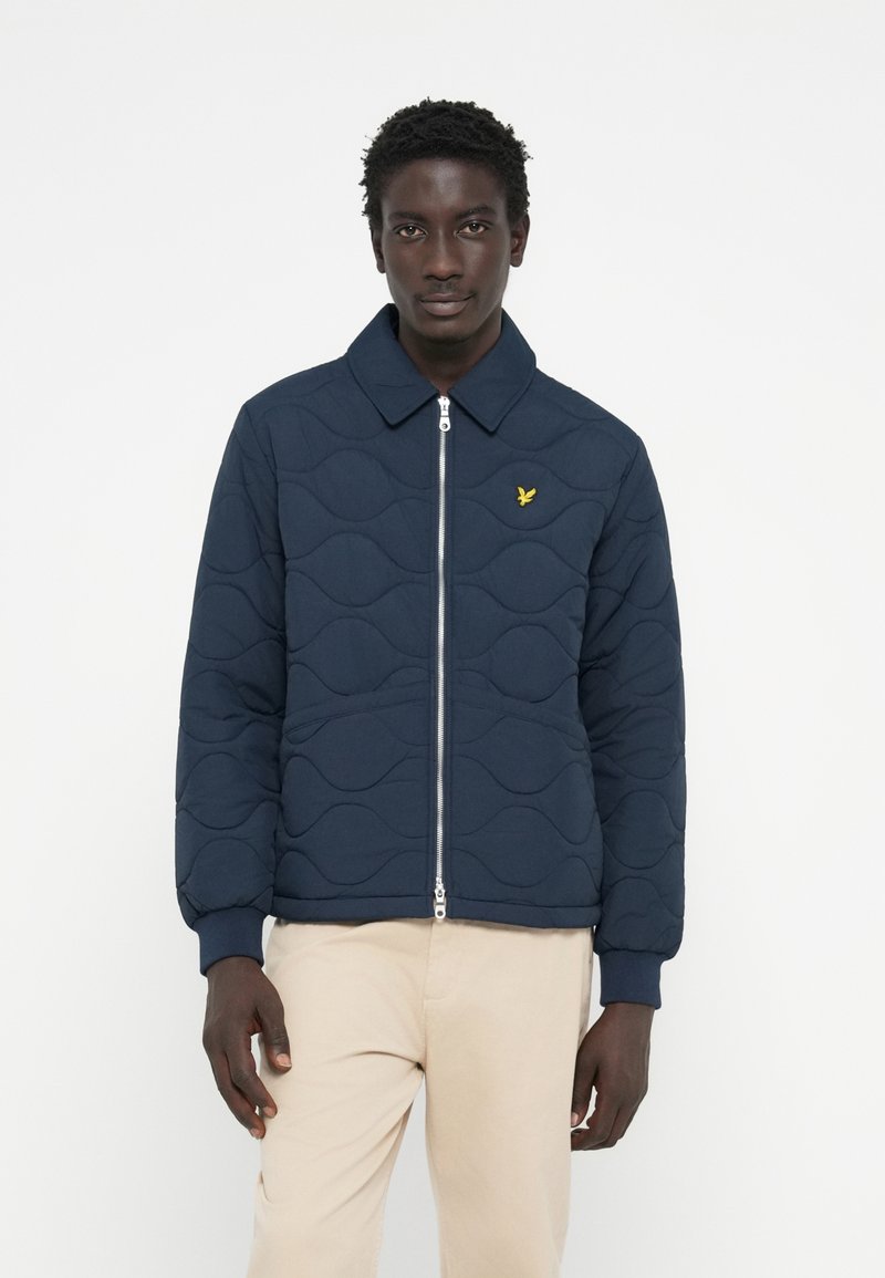 Marineblau gesteppte Jacke mit Reißverschluss, ausgestattet mit einem umgelegt Kragen und einem kleinen gelben Logo. Weiche Textur mit einem Wellenmuster auf dem Stoff.