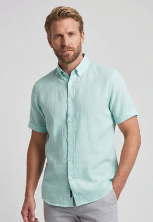 Blond man with beard wearing a light blue short-sleeve button-up shirt and light gray pants, standing with one hand in pocket.