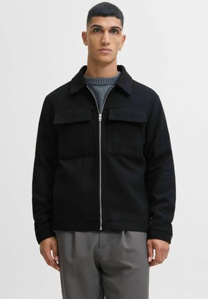 Schwarze Zip-Jacke aus strukturiertem Stoff mit Kragen, zwei Brusttaschen und taillierter Silhouette, kombiniert mit grauen Hosen.