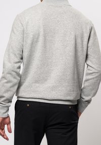 Grauer Pullover mit geripptem Kragen und Bündchen. Weicher, strukturierter Stoff und gerader Schnitt. Wird mit schwarzer Hose getragen, Rückansicht.