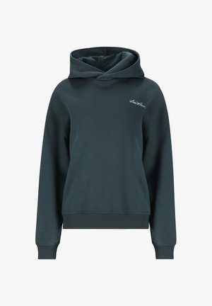 Jonge vrouw staand, gekleed in een donkerblauwe hoodie en bijpassende joggingbroek met witte sneakers, poseert met één hand in de zak tegen een witte achtergrond.