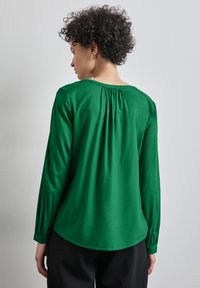 Blusa verde a maniche lunghe con vestibilità morbida, scollo rotondo, pieghe sul retro e polsini con bottone. Il tessuto sembra liscio e leggero.