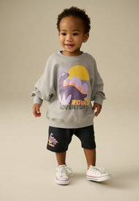 Sweatshirt gris avec un graphique de dinosaure, accents violets, texte "DINO ADVENTURE" ; shorts noirs avec un design de dinosaure similaire ; baskets blanches.