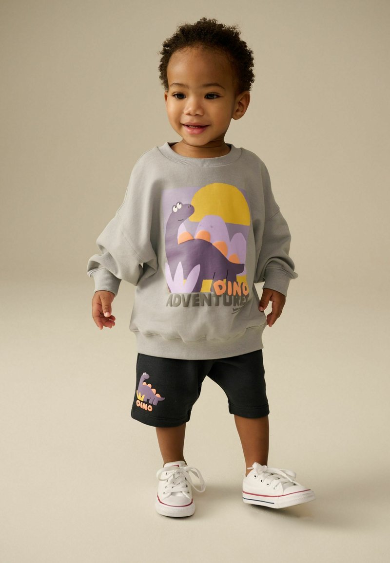 Sweatshirt gris avec un graphique de dinosaure, accents violets, texte "DINO ADVENTURE" ; shorts noirs avec un design de dinosaure similaire ; baskets blanches.