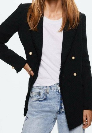 Kvinde iført en sort blazer med guldknapper over en hvid t-shirt og lyseblå højtaljede jeans med knaplukning, med den ene hånd på hoften.
