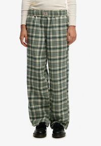 Pantalones de pijama a cuadros verdes y crema con un ajuste relajado, cinturilla elástica y textura de tela suave. Combinados con zapatos negros.