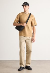 Ung mann iført en kremfarget kortermet skjorte, beige bukser, svarte joggesko og en svart caps, holder i stroppen til en svart crossover-veske.