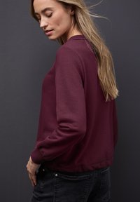 Sudadera burdeos con cuello redondo acanalado y puños. Tejido suave, corte relajado y dobladillo trasero ligeramente más largo. Llevada con vaqueros negros.