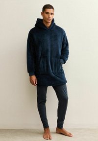 Man standing barefoot wearing a navy blue oversized hooded fleece sweatshirt and black fitted pants against a plain light wall.