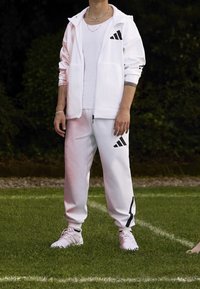 Bílý sportovní trenýrkový oblek s černým logem Adidas; zippová bunda a kalhoty na stažení, doplněné bílými sportovními botami; hladká textilie.