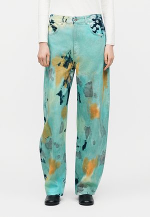 Τζιν παντελόνι με φαρδιά μπατζάκια και μοτίβα tie-dye σε τιρκουάζ, πορτοκαλί, μαύρο και γκρι, φορεμένο με μαύρα παπούτσια και λευκό μακρυμάνικο τοπ.