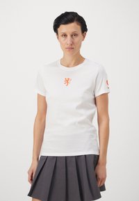 Hvit bomulls T-skjorte med et oransje løvegrafikk på brystet og en logo på ermet, sammen med en grå plisséskjørt.