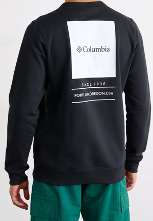 Man in een zwart Columbia sweatshirt met witte logo en tekst "Sinds 1938 Portland, Oregon, VS" op de rug, gecombineerd met groene cargobroek.