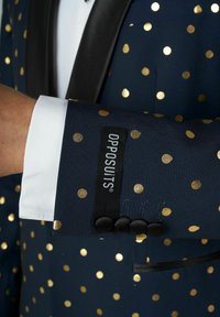 Námornícky blejzer s zlatými bodkami, saténovým čiernym klopom a bielymi manžetami. Obsahuje štítok "OPPOSUITS" a štyri čierne gombíky na rukáve.