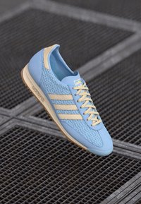 Zapatilla deportiva de color azul claro con parte superior texturada, rayas color crema y cordones. Presenta una suela beige y un diseño tejido en la punta.