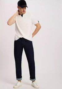 Biała koszula krótki rękaw z czarnym akcentem przy dekolcie, zestawiona z ciemnoniebieskimi dżinsami podwiniętymi na mankietach i białymi sneakersami.