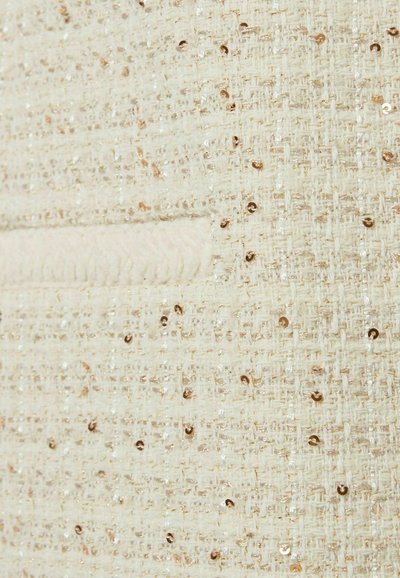Tissu crème texturé avec des paillettes dorées scintillantes intégrées. Présente une couture douce et surélevée courant parallèlement au bord.