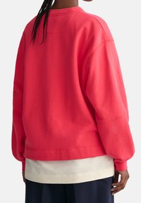 Koraliowy sweatshirt z długimi rękawami, luźny krój i okrągły dekolt. Nałożony na jasną koszulę, prezentowany z tyłu.