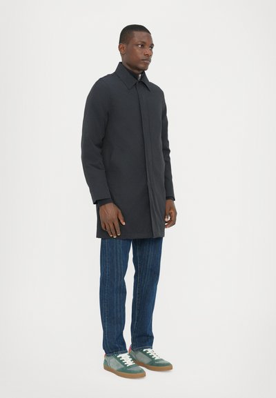 Zwarte jas met een hoge kraag, lange mouwen en een rechte snit, gecombineerd met blauwe textuurdenim en groene sneakers met witte accenten.