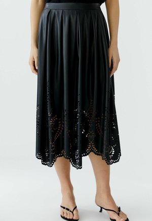 Frau trägt einen schwarzen plissierten Midirock mit laser-cut floralen Mustern entlang des wellenförmigen Saums, kombiniert mit schwarzen Riemchen-Pumps mit flachem Absatz.