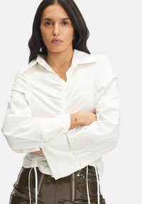 Blusa bianca di cotone con colletto, frontale arricciato e maniche ampie. Abbinata a una gonna verde brillante con cerniere.