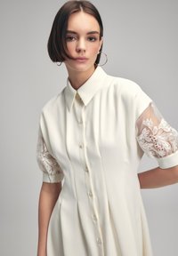 Witte shirtjurk met een kraag, korte puffmouwen in doorzichtig materiaal voorzien van ingewikkelde witte bloemenborduursel en een knoopsluiting aan de voorkant.