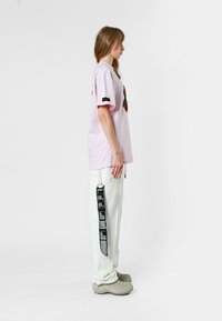 Camiseta rosa claro con un diseño bordado colorido, combinada con pantalones blancos que presentan detalles de etiqueta negra y zapatos beige gruesos.