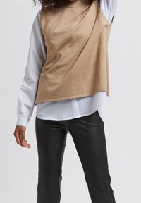 Beige asymmetrische top over een wit shirt, gecombineerd met zwarte aansluitende broek; gladde textuur, gelaagd ontwerp en contrasterende kleuren.