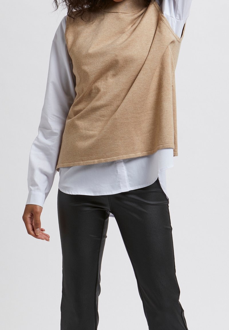 Beige asymmetrische top over een wit shirt, gecombineerd met zwarte aansluitende broek; gladde textuur, gelaagd ontwerp en contrasterende kleuren.