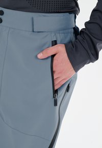 Grå, vattentäta byxor med en slät textur, med hög midja, fickor med dragkedja och kontrasterande svarta dragkedjor.