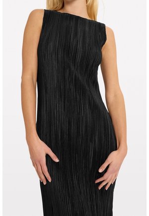 Sleeveless black dress with vertical pleats worn by a woman, hands resting on hips, neutral background.