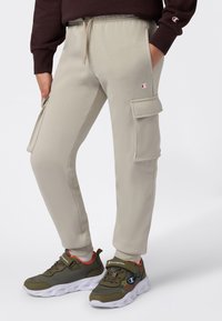 Beige cargo joggere med sidelommer og Champion-logoer, iført med olivengrønne og orange sneakers samt en mørkebrun sweatshirt.