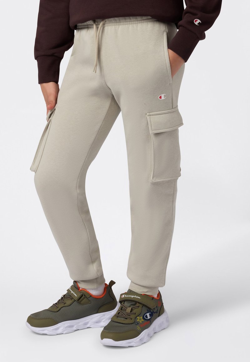 Beige cargo joggere med sidelommer og Champion-logoer, iført med olivengrønne og orange sneakers samt en mørkebrun sweatshirt.