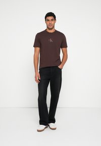 Καφέ κοντομάνικο T-shirt με μικρό λογότυπο, συνδυασμένο με μαύρο τζιν παντελόνι ίσιας γραμμής και λευκά αθλητικά παπούτσια με καφέ σόλες. Απλός σχεδιασμός.