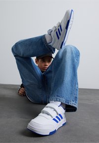 Białe buty sportowe z niebieskimi paskami, z solidną konstrukcją i rzepami. Noszone z luźnymi niebieskimi dżinsami. Szara teksturowana podłoga.