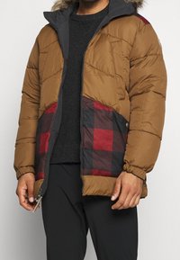 Uomo che indossa una giacca a piumino marrone con pannelli a quadri sulle tasche, un maglione nero sotto, pantaloni neri, e tiene la zip della giacca vicino alla vita.