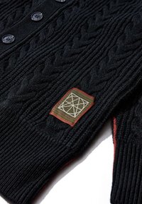 Cardigan nero lavorato a maglia con motivo a trecce, dotato di bottoni neri, una toppa testurizzata con un design geometrico e dettagli rossi lungo i bordi.