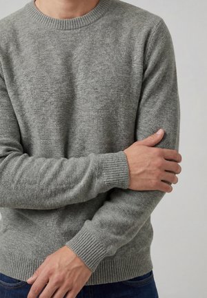 Uomo che indossa un maglione grigio aderente in maglia con polsini e orlo a coste, che tiene il braccio superiore sinistro con la mano destra contro uno sfondo semplice.